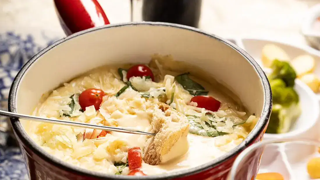 Mesa posta da Hannover com panela de fondue de queijo, vinho e acompanhamentos, com o garfo mergulhando o pão